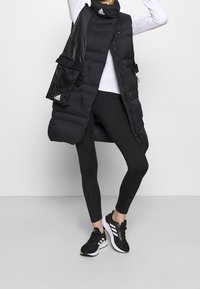 Sort oppustet vest med høj krave, lynlås foran, flere lommer, båret over en hvid langærmet top og sorte leggings, kombineret med sorte sneakers.