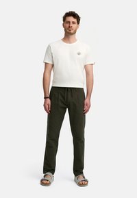 Hvid T-shirt med korte ærmer og logo, kombineret med løstsiddende, strukturerede mørkegrønne bukser. Sandaler med stropper fuldender outfittet.