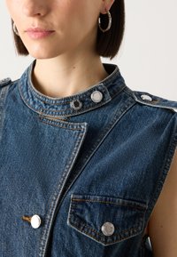 Denim prsluk s uzdignutim kragnom, bez rukava, dva džepa na prsima i srebrnim automatskim dugmadi. Tekstuirana plava tkanina s kontrastnim šavovima.