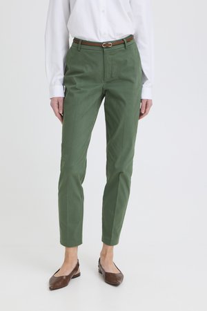 Femme portant un pantalon droit vert olive, une ceinture marron, une chemise blanche à manches longues et des ballerines marron pointues, debout sur un fond blanc.