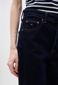 Σκούρο μπλε τζιν με πορτοκαλί ραφές, πέντε τσέπες και μικρή ετικέτα λογότυπου στην πίσω τσέπη. Μοντέλο με εφαρμοστή γραμμή και ψηλή μέση.