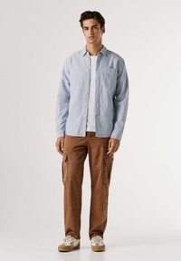 Camicia a righe azzurro chiaro e bianca con tasca sul petto. Pantaloni cargo marroni con tasche laterali. Sneakers beige con accenti azzurro chiaro.