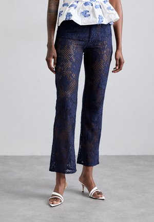 Pantaloni in pizzo di colore blu navy con una linea svasata. Presentano motivi intricati e una texture trasparente, abbinati a tacchi bianchi con cinturini.