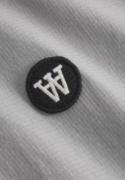 Un textile gris avec une texture ribbée subtile présente un patch circulaire noir. Le patch affiche des lettres blanches brodées "WW" dans une police stylisée.