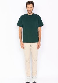 T-shirt in cotone verde scuro a maniche corte con colletto a girocollo, caratterizzato da un discreto logo del brand sul petto. Indossato con pantaloni beige chiaro e sneakers.