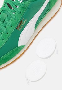 Zielony sneaker wykonany z tkaniny i zamszu z białym paskiem, z złotym logo, teksturowanymi sznurowadłami oraz dwoma białymi okrągłymi sznurowadłami obok.