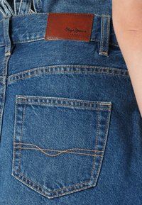 Farmernadrág hátsó zsebében egy barna bőrpamlaggal, amelyen a "Pepe Jeans London" felirat olvasható. Világoskék szín, látható varrási részletekkel.