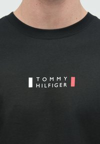 Black cotton t-shirt with a round neckline featuring the text "TOMMY HILFIGER" in white with red and white rectangular accents.