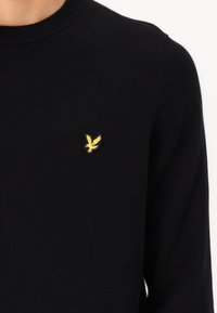Zwarte gebreide trui met een ronde halslijn en een klein geel vogellogo geborduurd op de linkerborst. Hengstige textuur, aansluitend ontwerp.