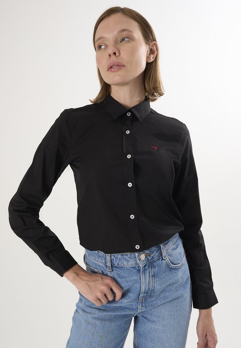 Sort skjorte med lange ærmer og klassisk krave, der har hvide knapper og et lille rødt logo på venstre side. Sammen med blå jeans.