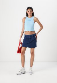Lys blå cropped tank top med "GAP" logo, kombinert med et denim mini skjørt, hvite høye sneakers og en rød veske. Enkelt design.