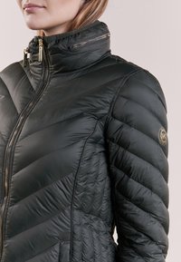 Sort puffjakke med høj krave, chevron-quiltet tekstur og guld lynlåse. Har et logo patch på venstre arm.