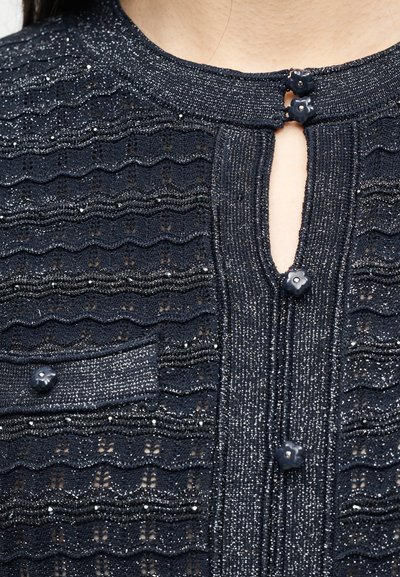 Κοντινό πλάνο σε μπλε σκούρο πλεκτό τοπ με υφή, ασημένιες κλωστές, κουμπιά με λουλούδια και μικρό άνοιγμα σε σχήμα κλειδιού στο ντεκολτέ.