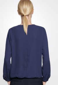 Blusa blu navy con maniche lunghe, texture liscia, dettaglio a chiave sulla schiena e polsini con bottoni. Design semplice ed elegante.