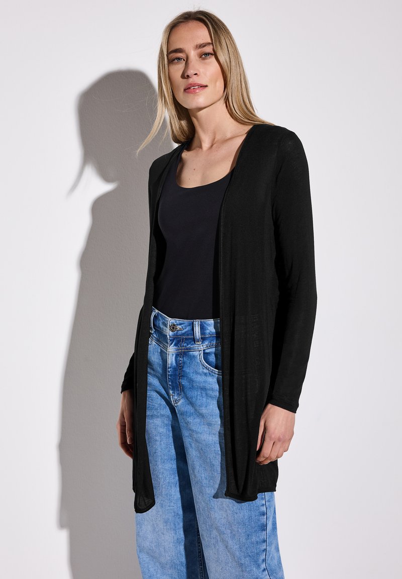 Svart lång kofta med öppen framsida, bärs över en svart tanktop och ljusblå, högmidjade jeans i denim, slät textur, minimalistisk design.