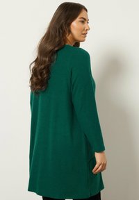 Zielony kardigan z długim rękawem wykonany z miękkiego materiału, o luźnym kroju, z bocznymi kieszeniami i dłuższym tyłem. Prosty design z czystymi liniami.