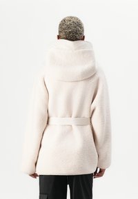 Light pink, fluffy fleece jacket with a large hood, belted waist, and long sleeves, seen from the back against a white background.