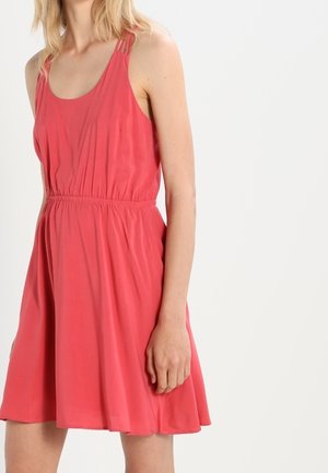 Kvinde iført ærmeløs koralfarvet kjole med samlet talje og plisseret nederdel, stående mod en ensfarvet hvid baggrund.