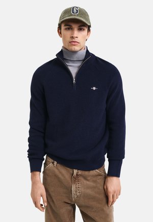 Navy Strickpullover mit halbem Reißverschlusskragen, gerippten Bündchen und Logo-Akzent. Getragen über einem hellgrauen Rollkragenpullover und kombiniert mit beigen Hosen.
