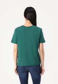 Smaragdgrønn bomulls t-skjorte med korte ermer, rundhals, og en avslappet passform, båret med mørkeblå jeans, sett fra baksiden.