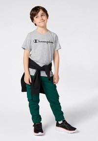 Pilkas medvilninis marškinėlis su „Champion“ logotipu, žali kargo kelnės su kišenėmis, juodas džemperis, rištas aplink liemenį, ir juodi sportbačiai su oranžiniais akcentais.