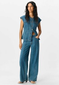 Chaleco de mezclilla con mangas en forma de capucha, cierre de botones y cortes laterales, combinado con pantalones de mezclilla de pierna ancha. Ambos presentan detalles de costura contrastante.