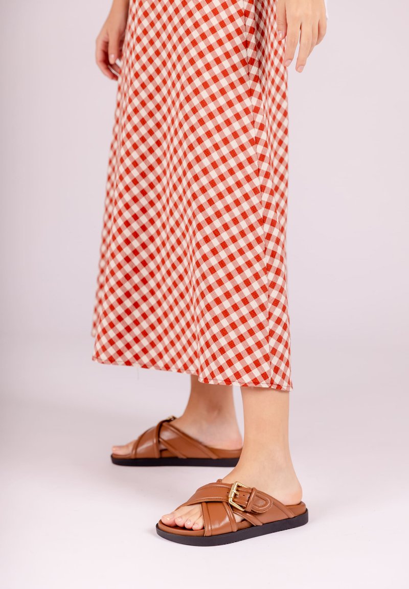 Bruine leren slip-sandalen met een brede band en een gouden gesp, gecombineerd met een rood-wit geruit lange rok. Soepele textuur, open teen ontwerp.