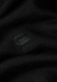 Tessuto a maglia nero con una trama fine, caratterizzato da un logo incorporato in una tonalità leggermente più scura. Il materiale appare morbido e flessibile.