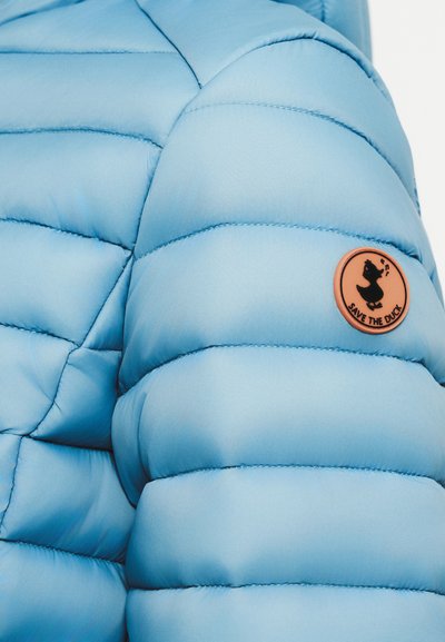 Veste matelassée bleu clair avec des sections matelassées, une texture lisse et un badge rond marron avec un logo de canard.