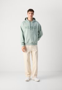 Giovane uomo in piedi che indossa una felpa mint green "HUGO", pantaloni crema e sneaker bianche, posando contro uno sfondo chiaro e semplice.