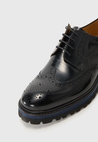 Sorte polerte skinnsko med brogue-detaljer, rund tå, grå snørebånd og en tykk såle med riller og blå aksentstripe.