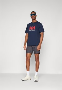 Tamno plavi pamučni majica s crvenim i bijelim logom "Helly Hansen", kombinirana s tamnim kratkim hlačama, svijetlim sportskim cipelama i bijelim čarapama.