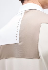 Οπίσθια όψη ενός ατόμου που φοράει μια μπεζ και λευκή μπλούζα με την επιγραφή "J.LINDEBERG" τυπωμένη κάθετα κοντά στον γιακά.