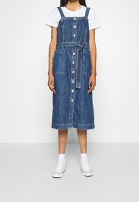 Dżinsowa midi sukienka z regulowanymi ramiączkami, paskiem i zapięciem na guziki z przodu. Posiada dwie przednie kieszenie i lekko wypłowiały niebieski odcień.