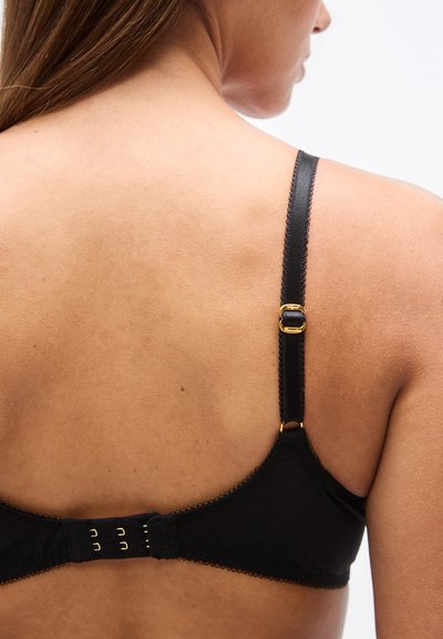 Μαύρο σουτιέν με κυματιστό περίγραμμα, ρυθμιζόμενες τιράντες και κούμπωμα με μεταλλικό στοιχείο σε χρυσαφί τόνο. Λείο ύφασμα με λεπτή υφή στο πίσω μέρος.