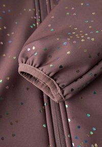 Prim-plan al unei mâneci din material mov pal, cu manșetă elastică și puncte mici irizate în formă de inimă împrăștiate pe țesătură.