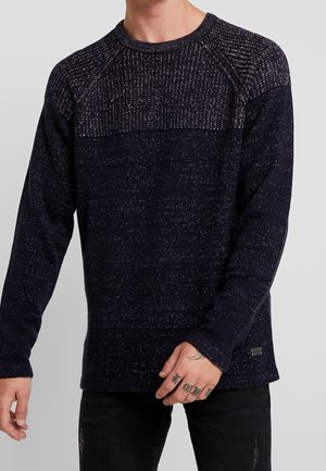 Mand iført en mørkeblå strikket sweater med tekstureret øvre bryst og skuldre, kombineret med sorte revnede jeans, tatovering synlig på hånden.