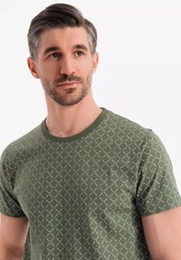 Zelené bavlněné tričko s geometrickým bílým vzorem, krátkými rukávy a kulatým výstřihem. Měkká textura, přiléhavý střih.