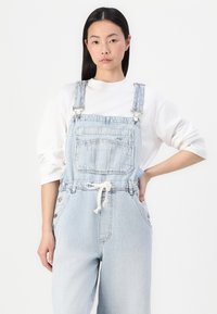 Des salopettes en denim bleu clair présentent des bretelles ajustables, une poche avant et une taille à cordon de serrage. Le tissu a une texture légèrement décolorée.