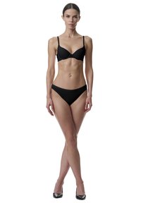 Vrouw die met gekruiste benen staat, gekleed in een zwarte bh, zwarte onderbroek en zwarte high heels tegen een witte achtergrond.