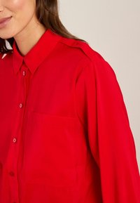 Rode button-up overhemd gemaakt van een gladde stof, met een borstzak en een klassieke kraag. De mouwen hebben een subtiele schouderdetail.