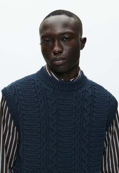 Sötétkék kötött ujjatlan pulóver, texturált kábelmintával, sötét csíkozott ing felett, klasszikus gallérral.