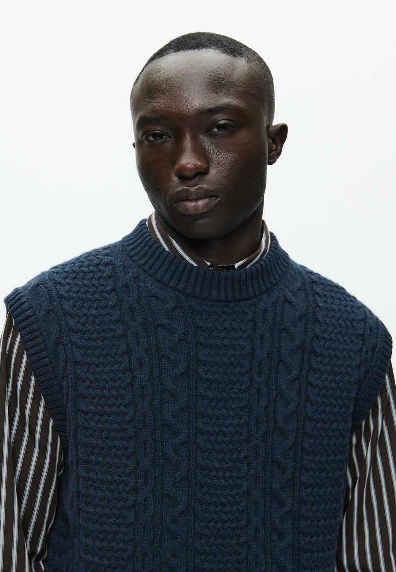 Pull sans manches en tricot marine avec un motif de câble texturé, porté sur une chemise à rayures foncées avec un col classique.