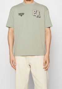 Jasnozielona bawełniana koszulka z prążkowanym okrągłym dekoltem, krótkimi rękawami oraz czarnymi haftowanymi logo "EA" i "Emporio Armani" na przodzie.