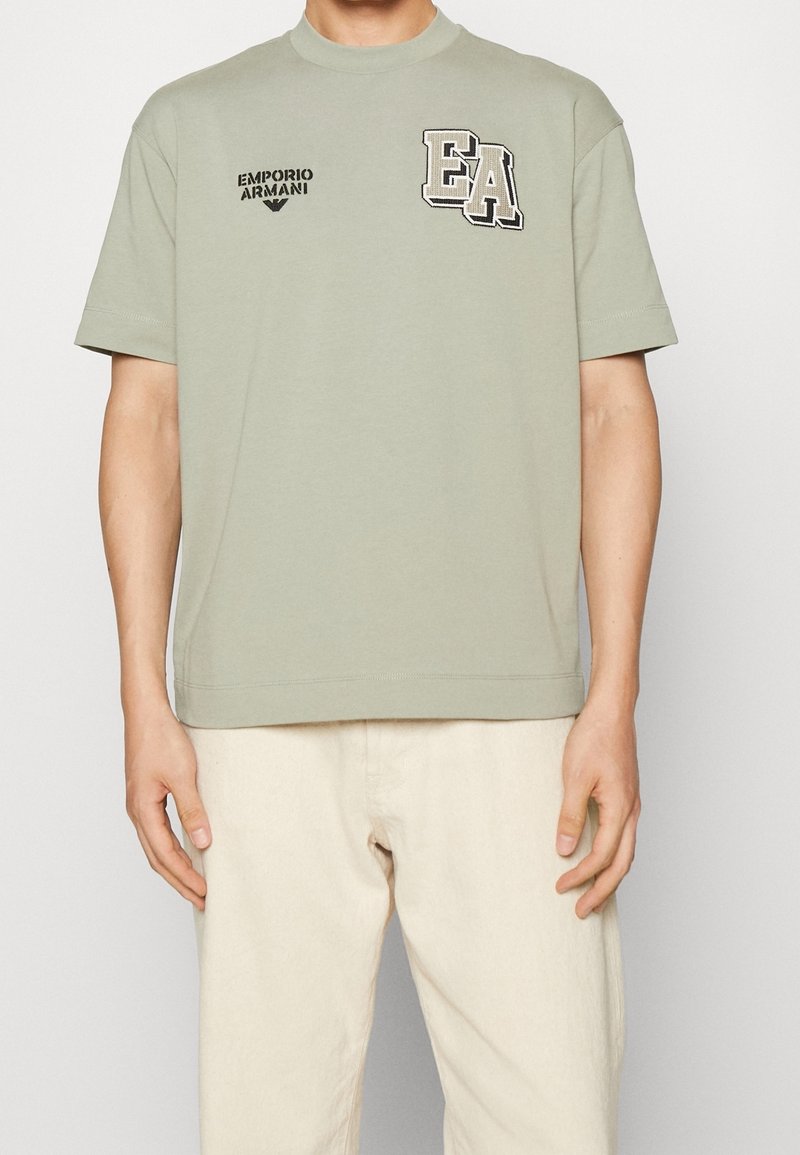 Jasnozielona bawełniana koszulka z prążkowanym okrągłym dekoltem, krótkimi rękawami oraz czarnymi haftowanymi logo "EA" i "Emporio Armani" na przodzie.