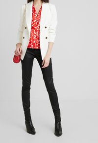 Blazer blanco con botones dorados, blusa de patrón rojo, pantalones negros ajustados y un pequeño bolso rojo de mimbre. Botas de tacón negro con puntera afilada completan el conjunto.