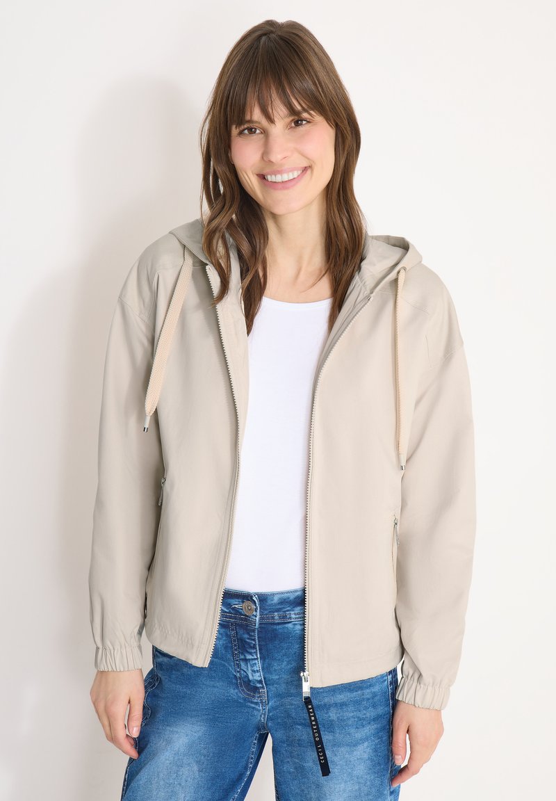 Femme souriante, portant une veste à capuche beige sur un chemisier blanc et un jean bleu, debout devant un fond clair uni.