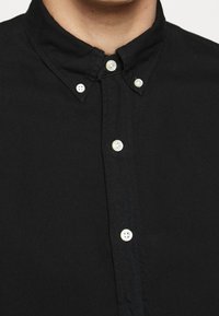 Svart skjorta med knappar i mjukt tyg, med klassisk krage och vita knappar, synliga sömmar längs placket.