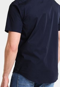 Koszula z krótkim rękawem w kolorze granatowym, wykonana z gładkiego materiału, z kołnierzykiem zapinanym na guziki i lekko zaokrąglonym dołem, noszona z dżinsami.
