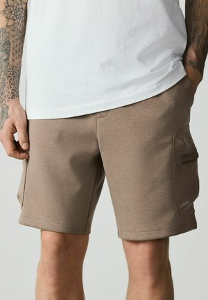 Mann trägt hellbraune Cargo-Shorts und weißes T-Shirt, mit sichtbaren Tattoos auf den Unterarmen, steht mit einer Hand in der Tasche.